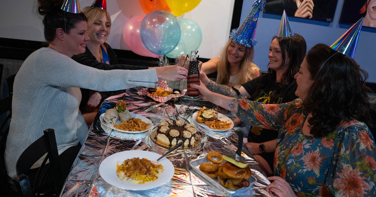 A group of patrons wearing festive birthday party hats toast over a table laden with food