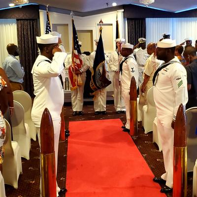 United States Navy ceremony with sailors saluting, red carpet, and flags.