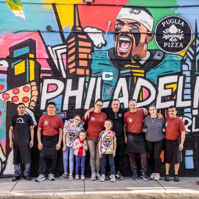 Restaurant staff and children posing in front of a Philadelphia-themed mural.