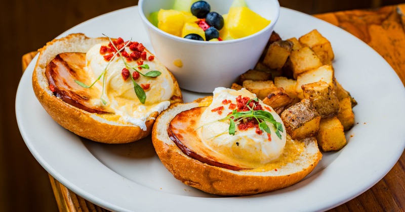 Eggs benedict served with diced potatoes and a small bowl of mixed fruit