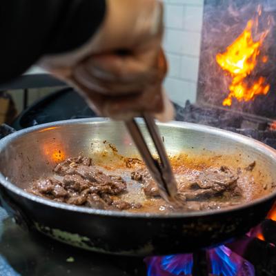 Chef cooking sliced beef in a hot skillet over an open flame.