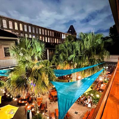 Patio at night with palm trees, teal shade sails, string lights, and picnic tables.