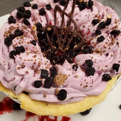 A purple cake with creamy swirls, topped with black berries and crumbled biscuits.