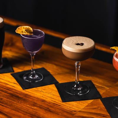 Espresso Martini and the purple cocktail placed on black cocktail napkins on a wooden counter.