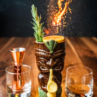 A Tiki mug filled with a cocktail, garnished with a sprig of rosemary and a citrus slice.