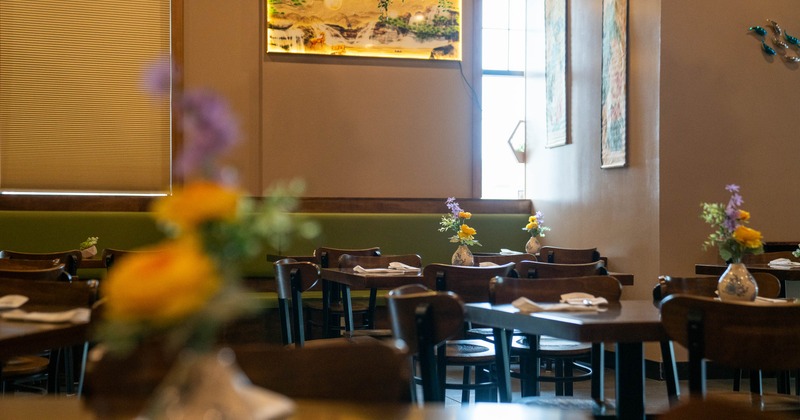 Warmly lit dining room - wooden tables, floral centerpieces, framed artwork, green bench seating