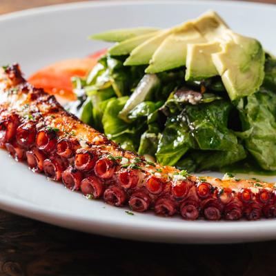 ZGrilled octopus served with cooked greens, avocado slices and tomato.