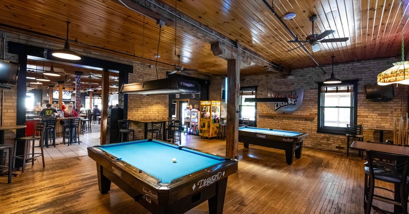 Interior, two pool tables and arcade machines, tables, chairs, wooden floors and brick walls