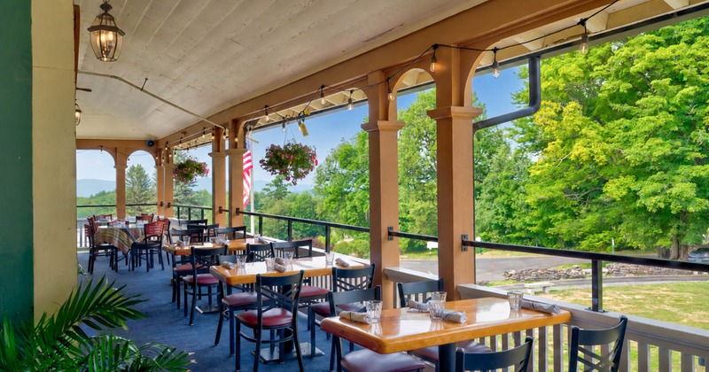 Outdoor dining area on a porch with wooden tables, black chairs, and hanging flower baskets
