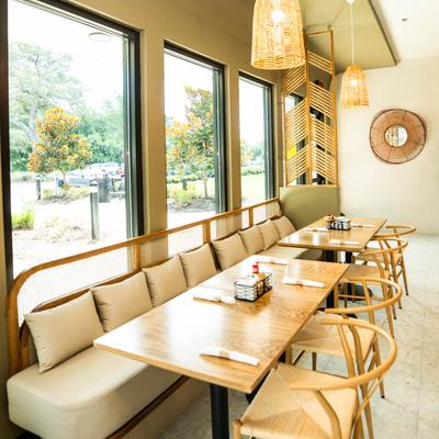 A tan cushioned bench with wooden tables and wicker chairs, arranged by large windows.