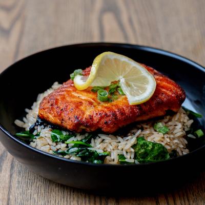 Grilled salmon served on a dark plate over rice with a lemon slice.