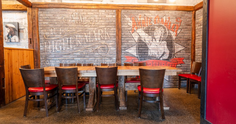 A long wooden table with six chairs set against a gray brick wall with murals and text