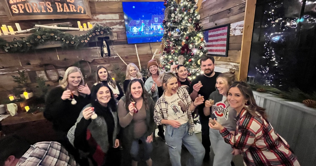 A group of smiling people pose in a cozy, festive setting with a Christmas tree and candles