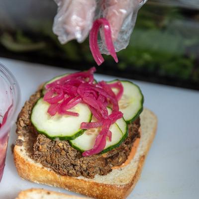 Sandwich with meat and sliced cucumbers being topped with pickled red onions.