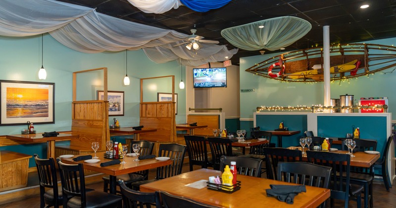 Interior with wooden tables, black chairs, blue-and-white ceiling drapery, and a decorative boat mounted on the back wall