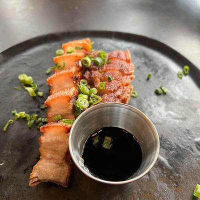 Pork belly garnished with  green onions on a black plate, accompanied with a dark dipping sauce.