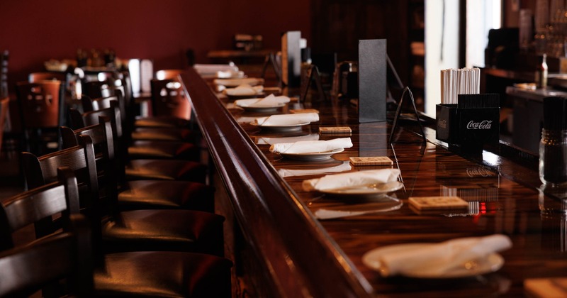 A view of a restaurant bar featuring a polished wooden counter with white napkins and plates