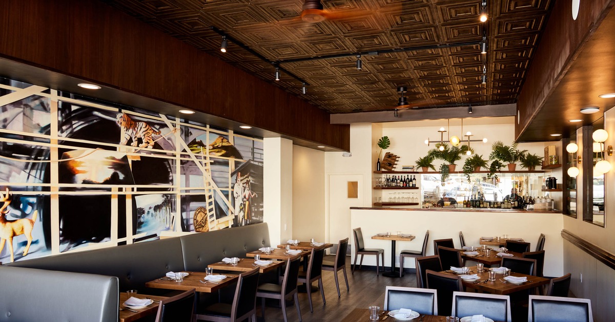 Interior of a modern restaurant with a mural, tables, chairs, and a bar area decorated with plants