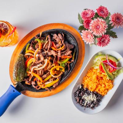 Sizzling steak fajitas served beside pink flowers with rice, beans, and a cocktail.
