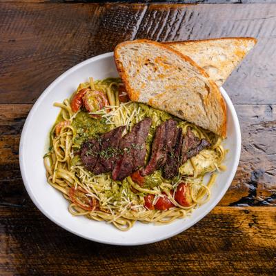 Sliced grilled steak served over green pesto pasta with tomatoes and toasted bread.