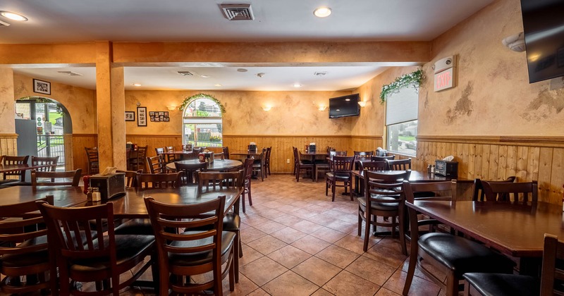 Interior of a restaurant with wooden tables and chairs, arched windows, and wall-mounted television