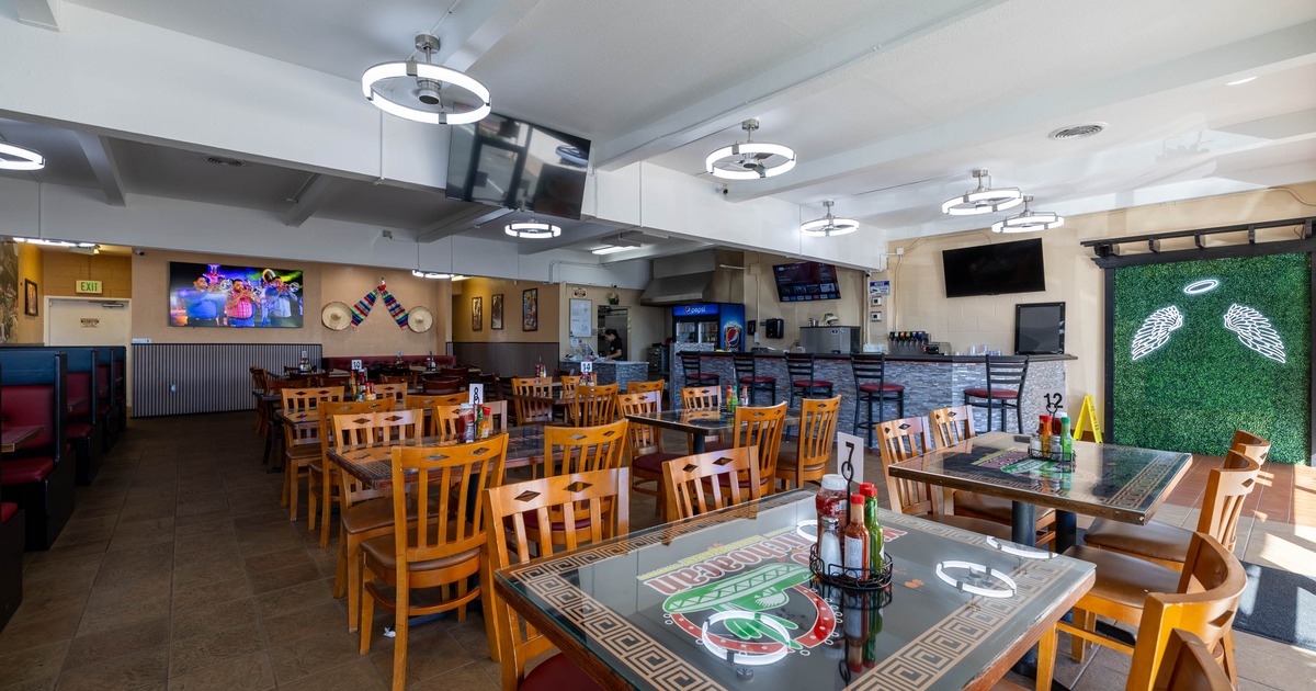 A  brightly lit interior - wooden tables, chairs, booth seating, TVs, a hedge wall with angel wings