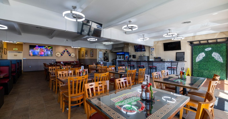 A  brightly lit interior - wooden tables, chairs, booth seating, TVs, a hedge wall with angel wings