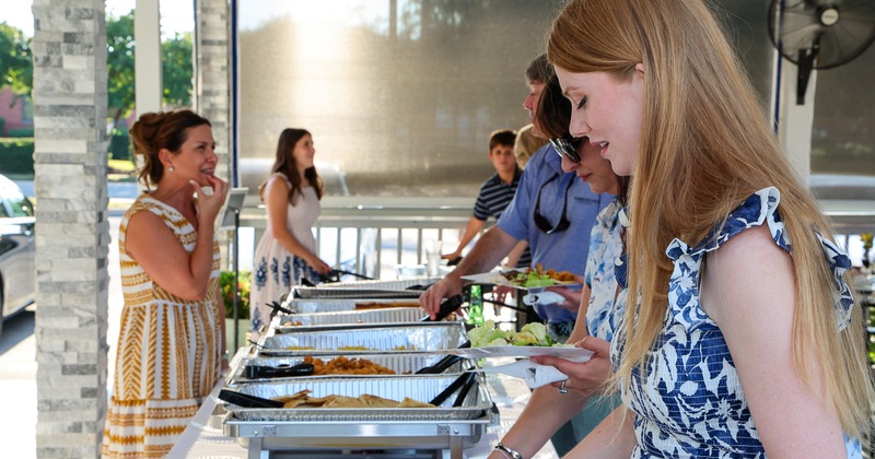 People gathered around a buffet setup on a covered patio during a daytime event.