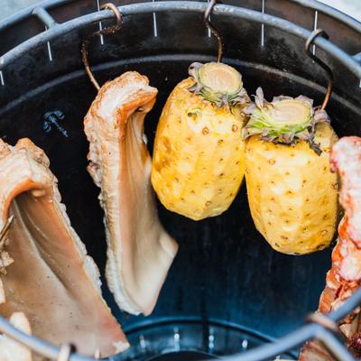 Barrel smoker with whole pineapples and pieces of meat hanging vertically.