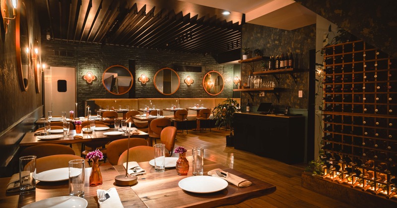 Interior with wooden tables, warm lighting, a wine rack, and circular mirrors on dark brick walls