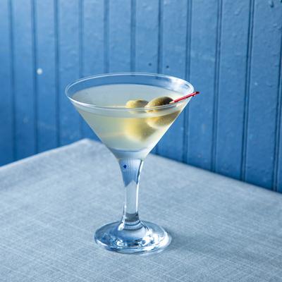 A martini, garnished with olives, sits on a gray tablecloth in front of a blue wooden wall.