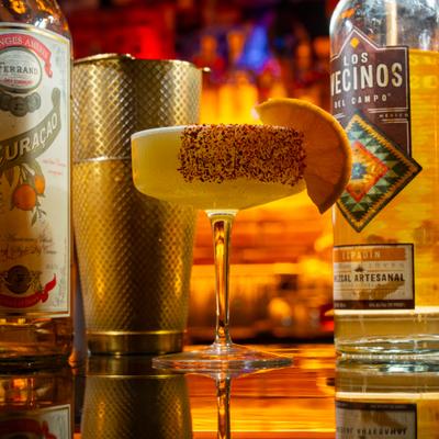 Roasted Pina Rita cocktail sits on a reflective bar top beside a shaker and a mezcal bottle.