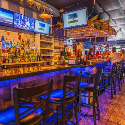 Sport bar interior with a long counter, blue lights, TVs, and shelves of liquor.