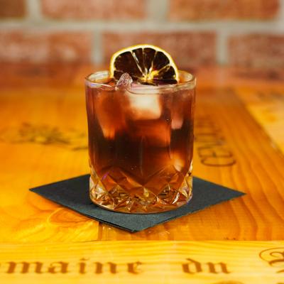 Dark cocktail over ice garnished with dry citrus, served on a wooden table.