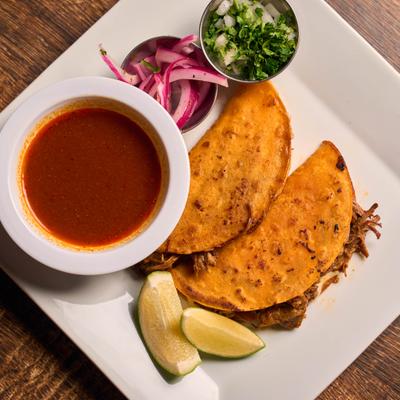 Quesabirria tacos served with dipping broth, chopped cilantro, onions, and lemon wedges.