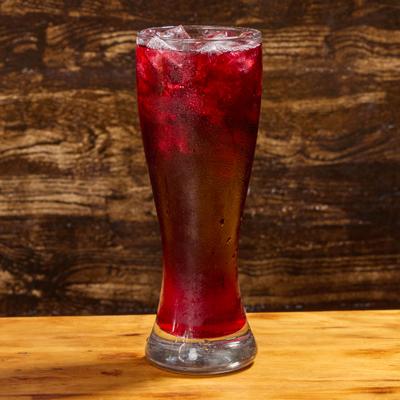 A tall glass of Jamaica drink, sits on a light wooden table against a textured brown background.