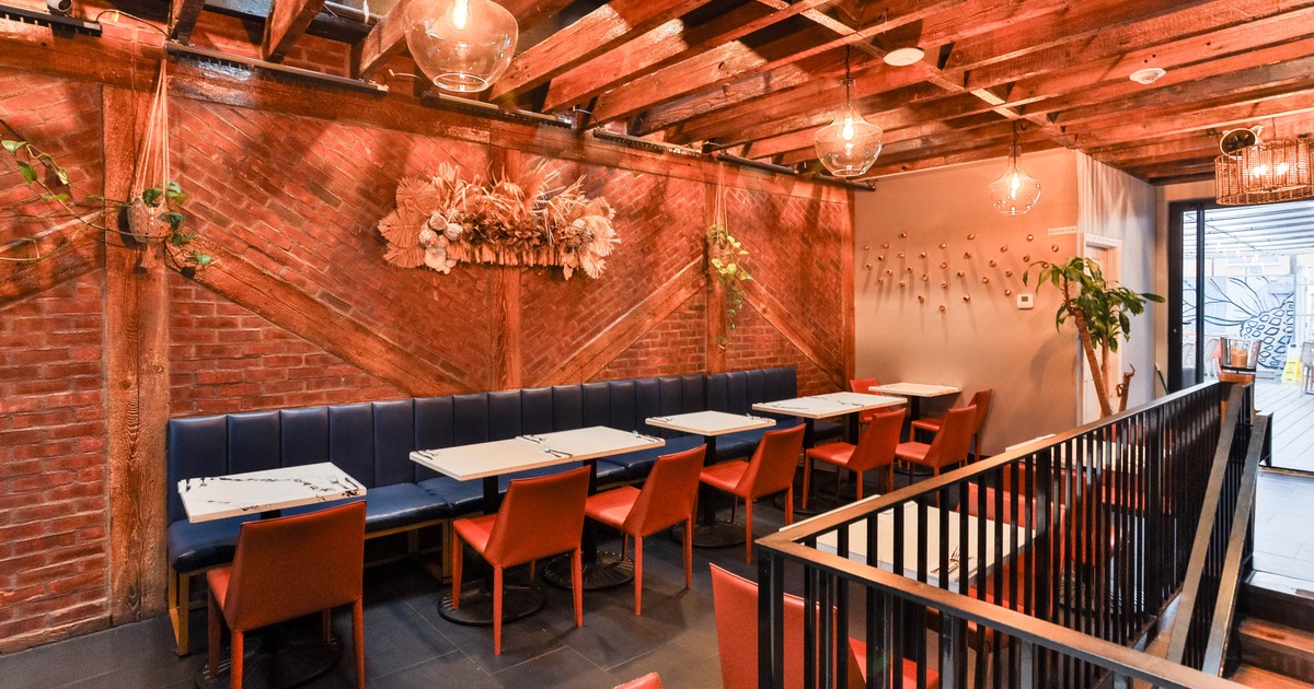 Restaurant interior with brick walls, dark wooden beams, blue banquettes, red chairs, and marble-like tables.