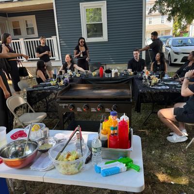 People at an outdoor gathering around a table and a grill.