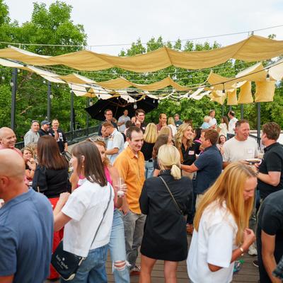 Crowd socializing on outdoor patio under fabric canopies and string lights.