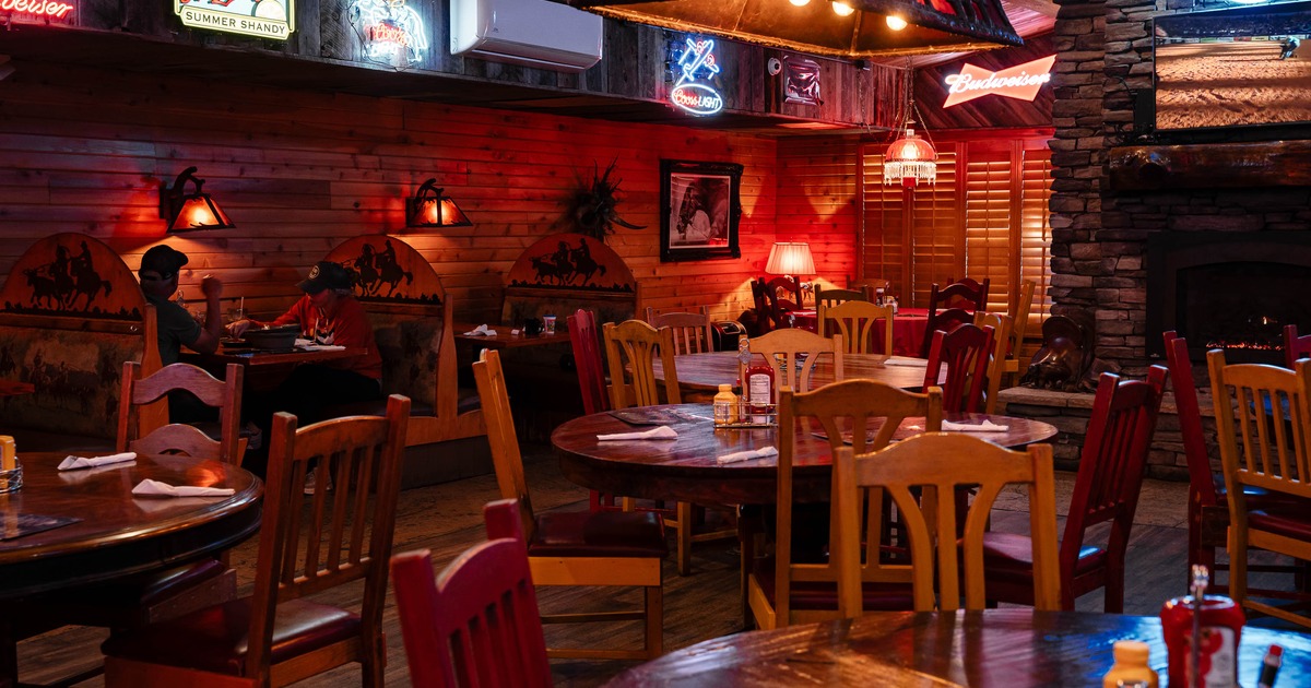 Rustic restaurant interior with wooden tables, chairs, neon beer signs, and a stone fireplace