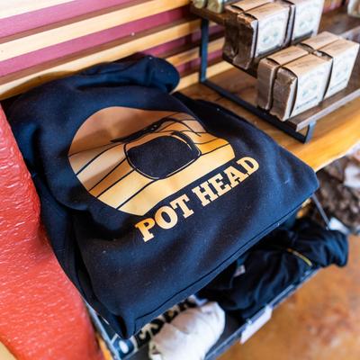 A black hoodie with a golden POT HEAD graphic is displayed on a shelf next to packaged goods.