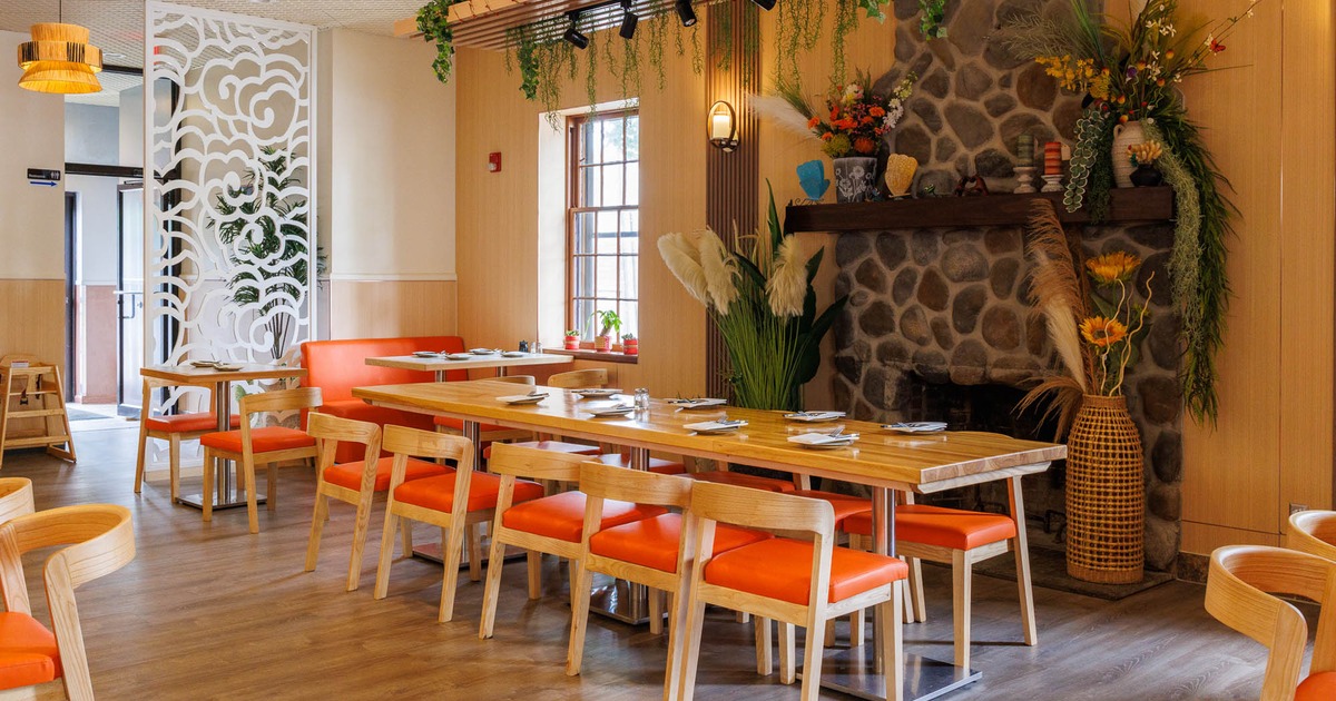 Restaurant interior, wooden furnishings, orange cushions, a stone fireplace, and decorative plants.