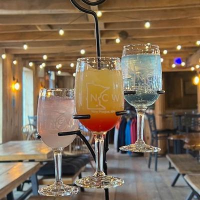 Three colorful drinks in glasses on a metal stand, set in a cozy wooden interior with string lights.