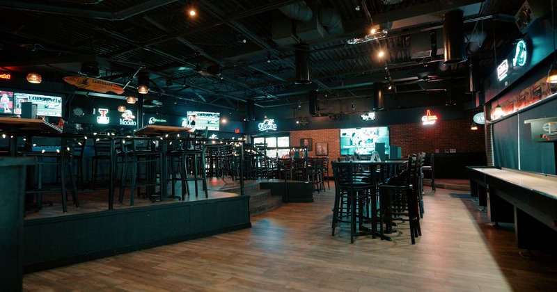 Interior of a dimly lit bar with wooden floors, high tables, neon signs