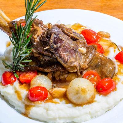 Braised meat rests on creamy mashed potatoes with tomatoes and pearl onions.