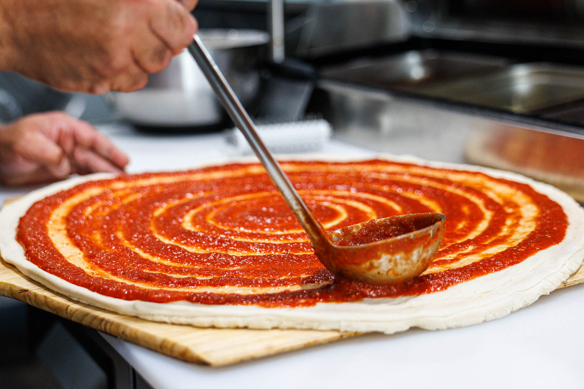 Pizza maker spreads tomato sauce on pizza dough using a ladle