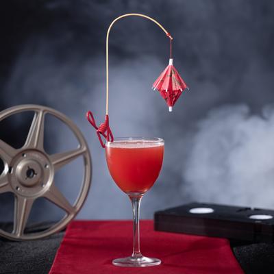 A red Kanpai cocktail with a paper umbrella garnish sits beside a film reel and VHS tape.