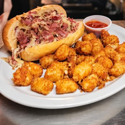 A Reuben sandwich served with tater tots and dipping sauce on a white plate..