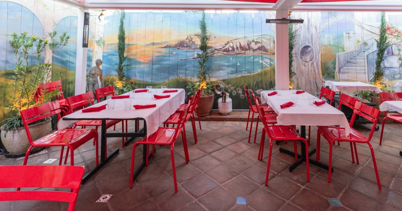 Outdoor dining area with red chairs, white tablecloth-covered tables, and a scenic mural on the wall