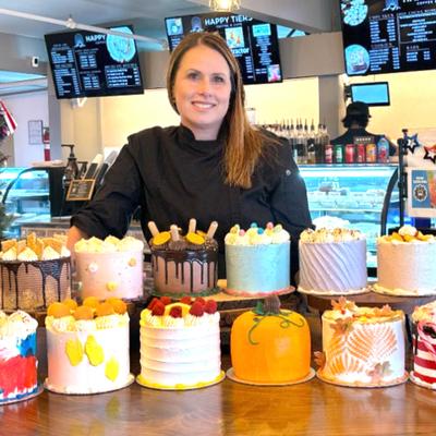 A baker stands behind an array of decorated cakes on a wooden counter.